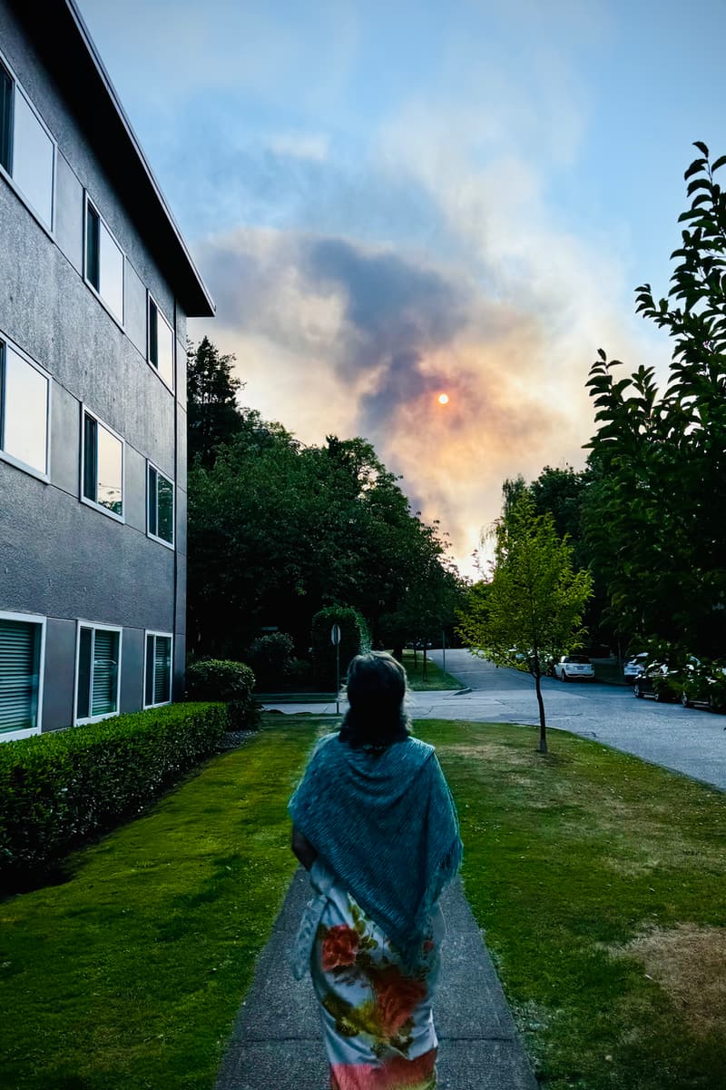 Photo of a pillar of black smoke on the horizon, looking down a residential street with an apartment building on the left and trees and street on the right. There’s a woman in the foreground walking towards the smoke, looking up at the sky.