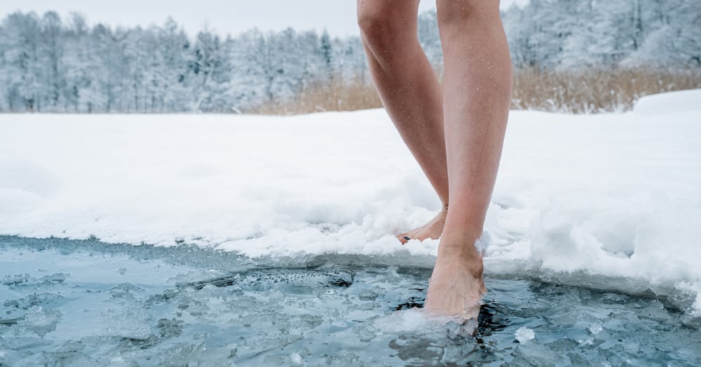 Photo of a winter landscape. In the foreground, a woman’s legs and an icy lake: one foot is on the snow, and the other is stepping into an extremely icy pond or stream.
