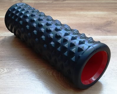 Photo of a massage roller on a hardwood floor. It’s a hollow tube with a black foam rubber coating and many prominent knobblies. It’s surface has the texture of a ATV tire.