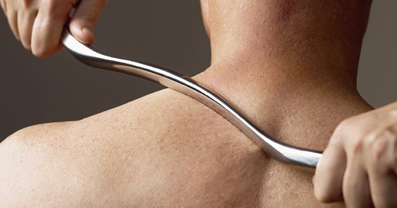 Photo of a Graston technique, a massage modality, being applied man’s upper back and shoulders, using a chrome tool about a foot long, slightly curved.