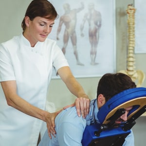Photo of a female massage therapist in a lab coat massaging a male client in a clinical environment, with charts and a spine model in the background.