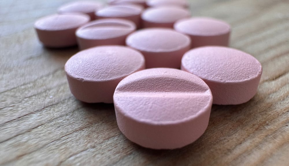 Photo of a group of pink tablets scattered on a wooden surface. The drug is rosuvastatin, but there’s no obvious indication of this. The tablets are round and appear to be uniform in size and shape. Some of them are placed with the imprinted side facing up, showing a line across the center, which suggests they can be split in half easily. The focus is sharpest at the tablet in the foreground and gradually softens toward the back, giving a sense of depth. The lighting suggests an indoor setting with natural light, which casts soft shadows behind each pill.