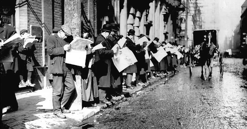 Vintage photo, probably pre-1920, showing a lot of people reading newspapers while standing on a snowy and icy sidewalk, with a horse-drawn carriage passing by. The man in the foreground is leaning against a telephone pole as he reads, and his head is tilted well forward, about a 45˚ angle. Several other people in background have the same posture.