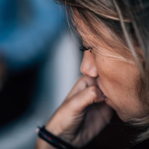 A woman with blonde hair looks down pensively, hand raised to her mouth, with a blurred figure in blue in the background — suggesting a therapy or counselling session.