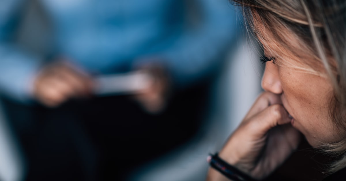 A woman with blonde hair looks down pensively, hand raised to her mouth, with a blurred figure in blue in the background — suggesting a therapy or counselling session.
