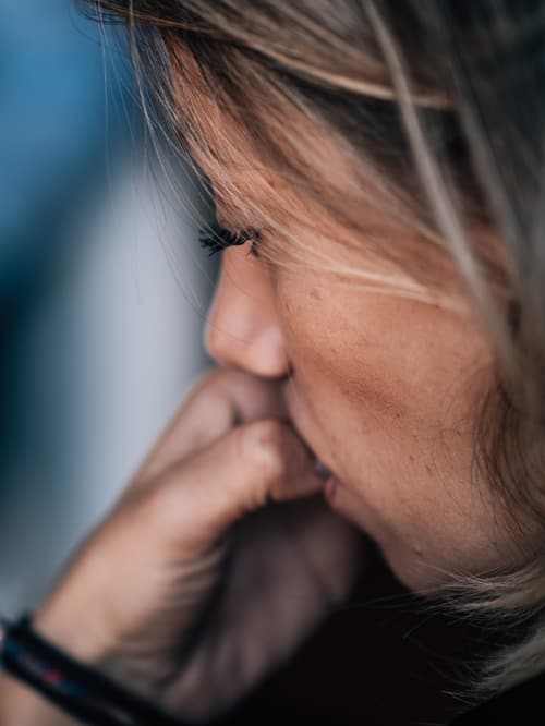 A woman with blonde hair looks down pensively, hand raised to her mouth, with a blurred figure in blue in the background — suggesting a therapy or counselling session.