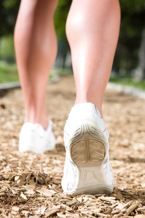 Stock photo of a close-up of a person’s lower legs and feet and white shoes as they walk on a trail covered with wood chips. The background is blurred but appears bright and green, and the setting is probably a park.