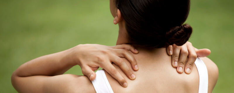 Image representing trigger point pain. Very wide format photo of the back of a woman’s head and bare shoulders, in front of an out of focus green background. We can’t see her expression, but she’s looking up, and grasping both shoulders with both hands — apparently she has some pain, perhaps myofascial trigger points in her shoulders.