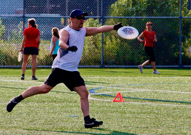 A man wearing a sleeveless athletic shirt, shorts, gloves, sunglasses, and a cap is lunging forward on an artificial turf field while extending his right arm to catch or throw a white flying disc.
