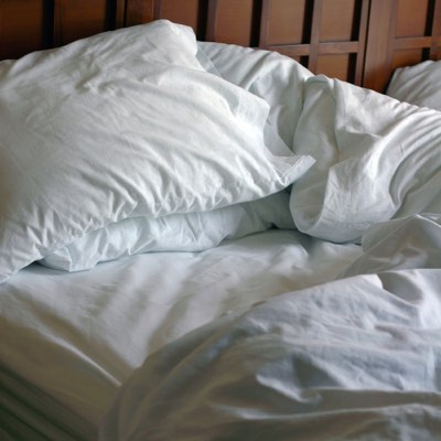 Photograph of an unmade bed with rumpled pillows in the foreground, representing the relationship between pillows, sleep, and neck pain.