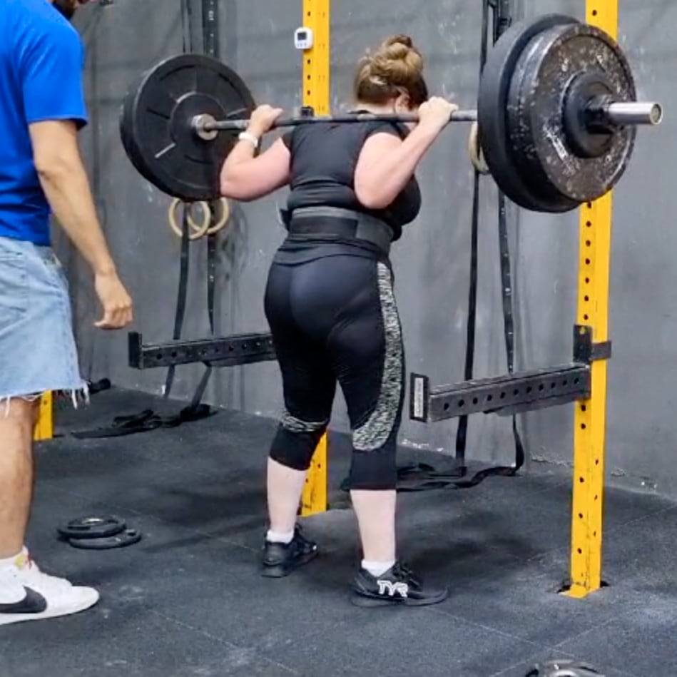 Photo of a woman, Cara Bereck Levy, lifting a heavy barbell — specifically a back squat with a loaded barbell in a gym. She is standing in a squat rack, holding the barbell across her upper back while wearing a lifting belt for support. She is dressed in a black top, black leggings with patterned side panels, and black shoes. A coach or spotter in a blue shirt and jean shorts is standing nearby, monitoring her form. The squat rack is bright yellow, and the gym has a rugged, industrial feel with gymnastic rings and weight plates visible in the background.