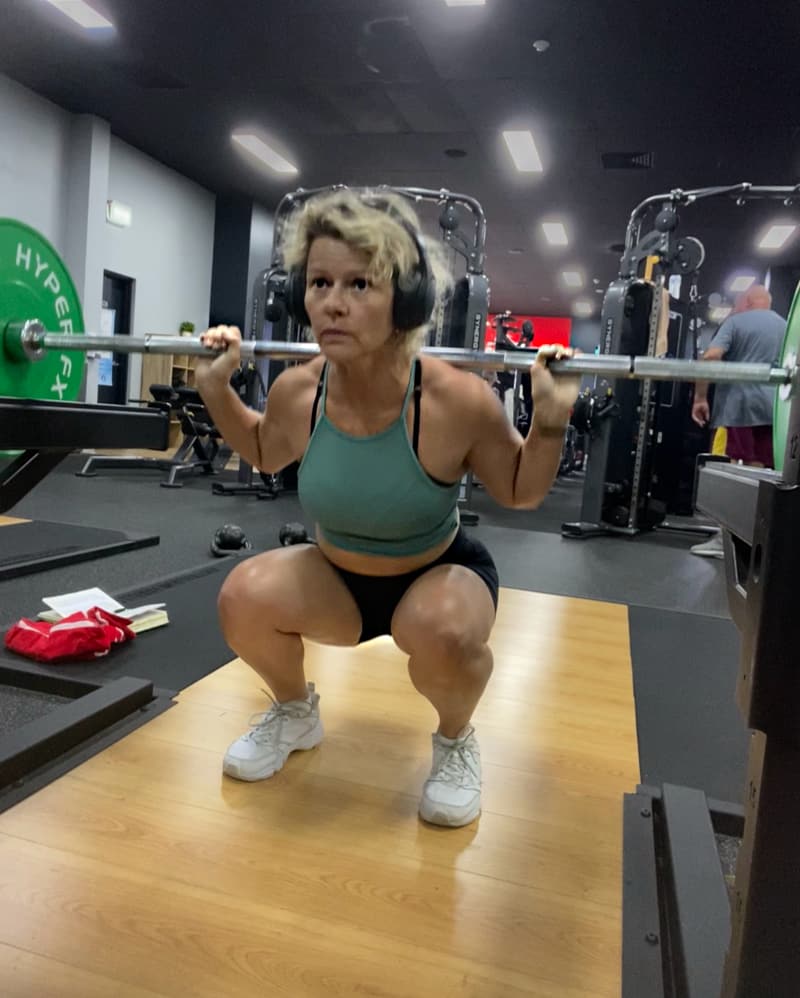 Photo of a woman, Kelly Lobgeier, in the low position of a back squat in a gym. She is holding a barbell across her shoulders, with at least 50lbs of plates on it, possibly more that aren’t visible. She’s wearing large headphones, a green sports bra, black shorts, and white athletic shoes. She is squatting on a wooden platform with green weight plates on the bar. Her expression is focused as she performs the lift. The gym background includes various strength training machines and equipment, along with a few other people.