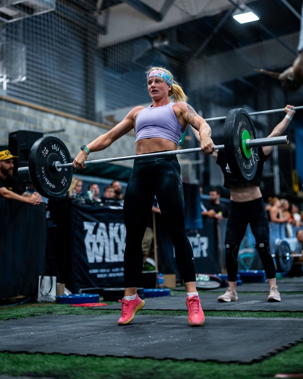 Photo of a woman, Lois Smith, lifting a 30kg on a barbell, up on her toes mid-snatch, almost jumping, during a weightlifting or CrossFit competition. The barbell at hip height, as she pulls the bar upward with intense focus. She is wearing a lavender sports bra, black leggings, and bright pink athletic shoes. A colourful headband keeps her hair in place. In the background, other athletes and spectators can be seen in a large gym or indoor facility with high ceilings and equipment. The scene is full of energy and focus as she competes.