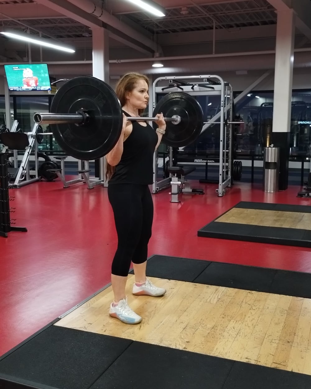 Photo of a woman, Mariana Molinari, holding a heavy barbell at chest height in a gym. The barbell is in the front rack position, preparing for a lift. She is wearing a black tank top, black capri leggings, and white athletic shoes with pastel accents. She appears focused, maintaining an upright posture as she holds the barbell across her shoulders. The gym background features weightlifting platforms, machines, and a television playing in the upper left corner.