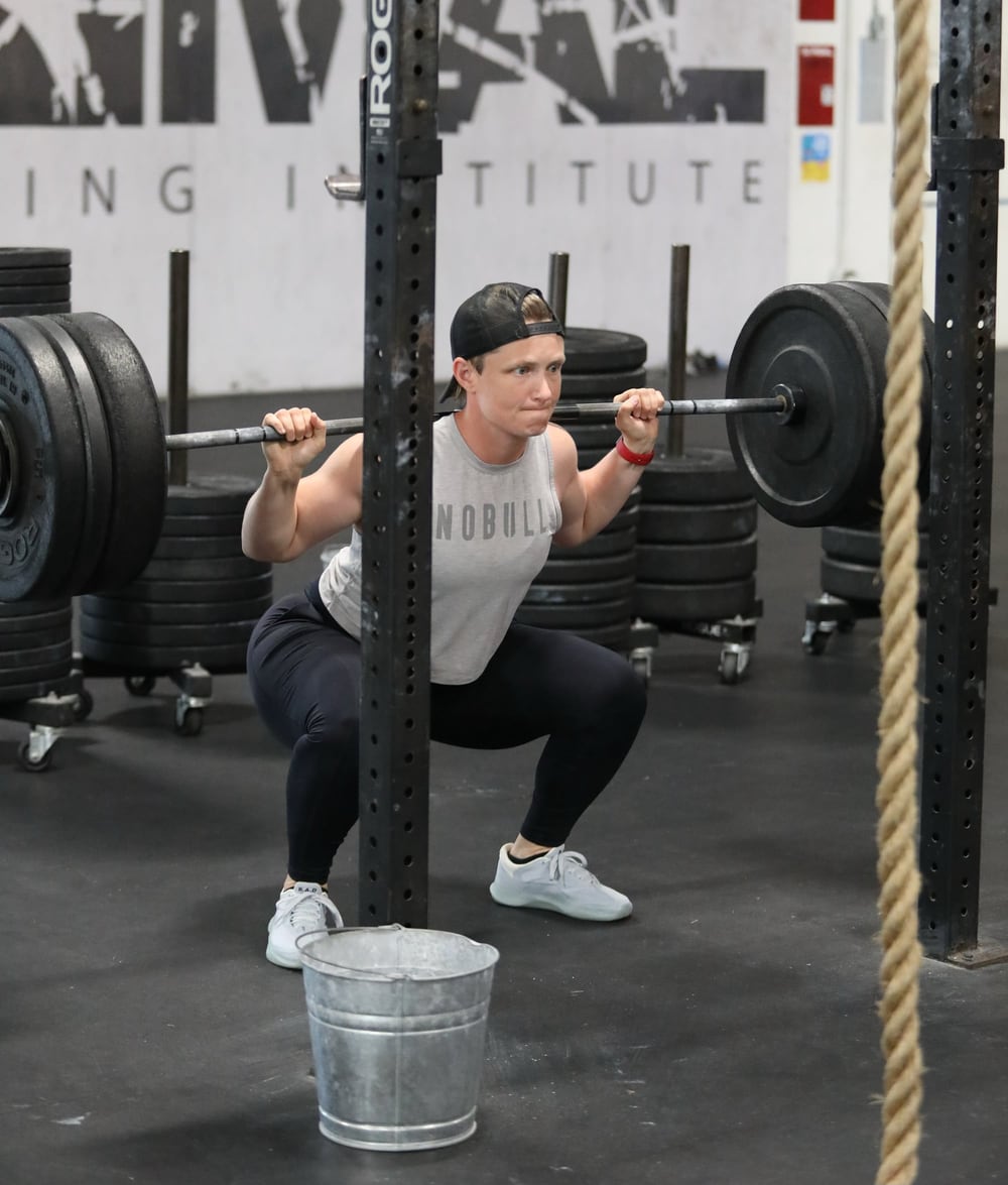 Photo of a woman, Rachel Moore, in a deep squat with a very heavy barbell on her shoulders, probably about 90 kg or 200 lbs. She is wearing a gray “NOBULL” tank top, black leggings, white athletic shoes, and a backward black cap. The gym features stacks of weight plates, a squat rack, and a rope hanging in the background. A small metal bucket is positioned in front of her, likely part of her setup or training environment. Her expression shows focus and determination during the lift.