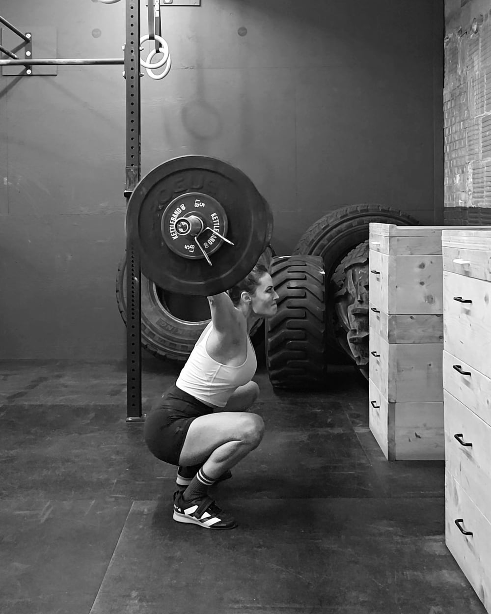 High quality B&W photo of a woman, Stephanie Fraser, performing an overhead squat in a gym. She is holding a barbell overhead with fully extended arms, squatting deeply with her hips below her knees. She is wearing a white tank top, black shorts, and weightlifting shoes with high socks. In the background, there are large tires and wooden plyometric boxes, indicating a functional training space. The gym setting is minimalist, and her expression shows focus as she maintains stability and strength in the squat position.