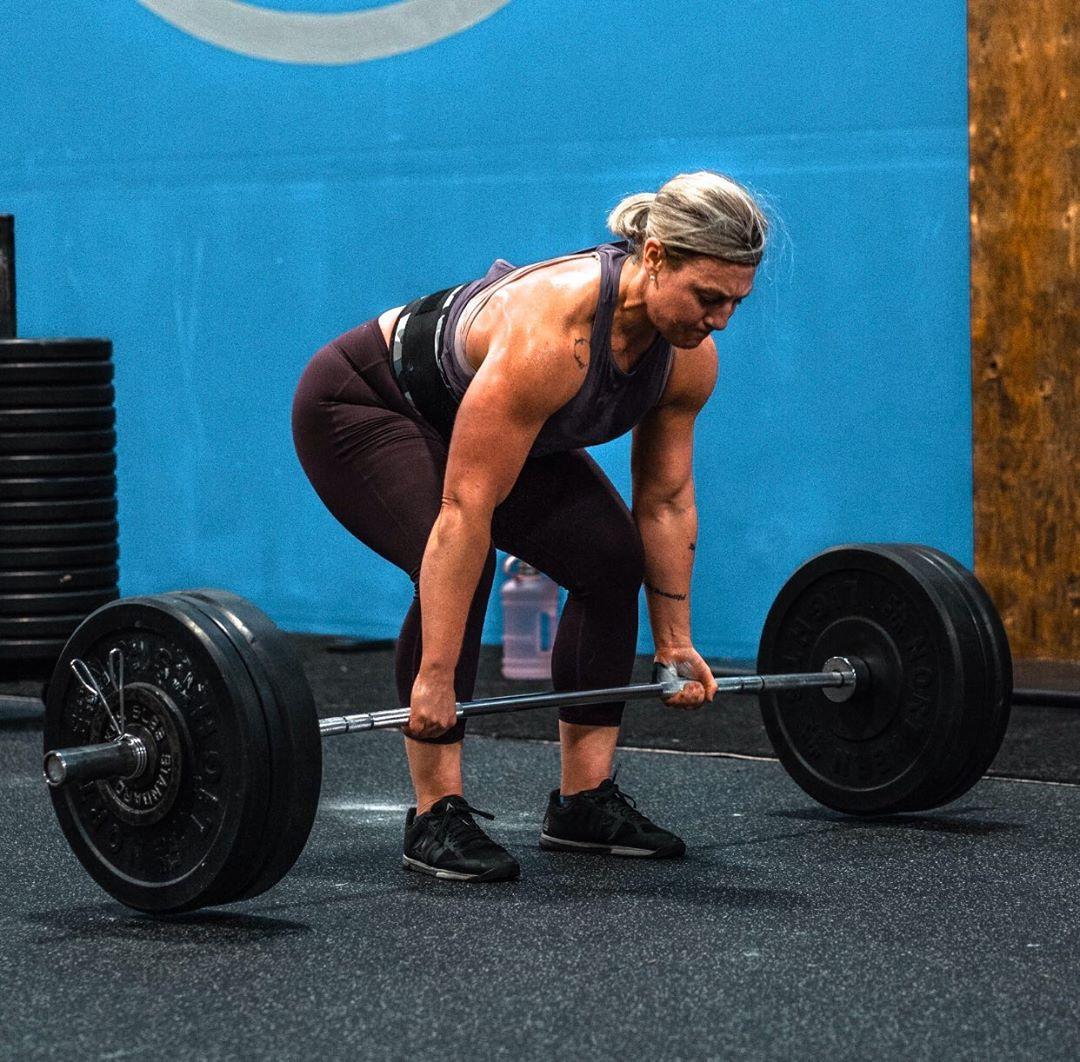 Photo of a woman, Tara Hawkin, at the beginning of a deadlift of a heavy barbell, probably about 90 kg or 200 lbs. She is bending over with a neutral spine, gripping the barbell on the floor. She is wearing a dark tank top, maroon leggings, black athletic shoes, and a lifting belt for support. Her expression is focused as she prepares to lift the barbell. The background features a blue wall and some stacked weight plates.