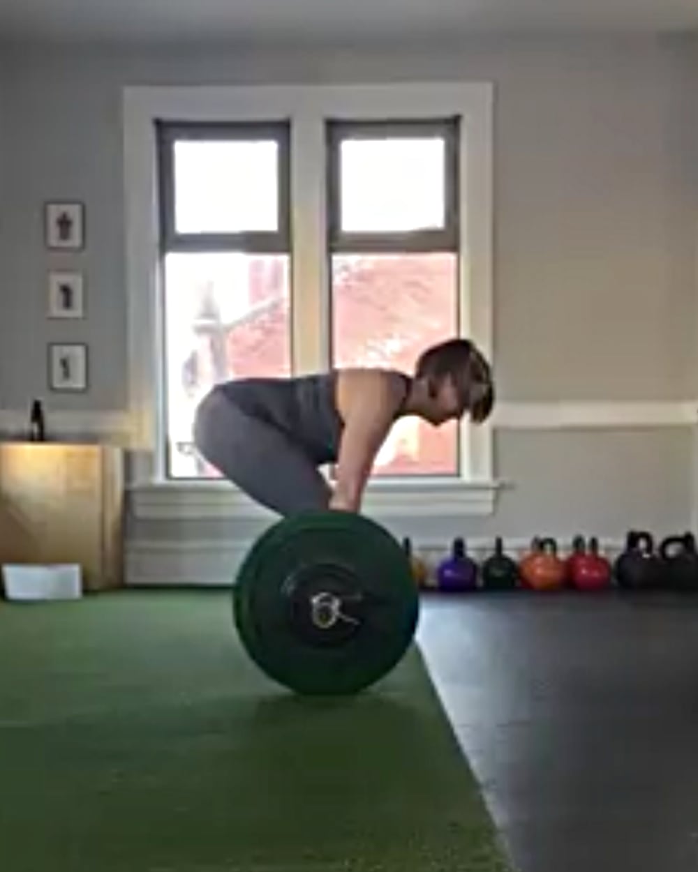 Photo of a woman, Terri Rowan, performing a deadlift in a home gym, showing her exactly side on from a distance of a few metres. She is stooped forward with a perfectly horizontal back, holding a barbell on the ground with green weight plates, preparing to lift it. She is dressed in a fitted, sleeveless gray top and matching leggings. The room has large windows in the background, letting in natural light, and several kettlebells are lined up along the wall. The space appears calm and organized, with a green workout mat under the woman as she focuses on her lifting form.