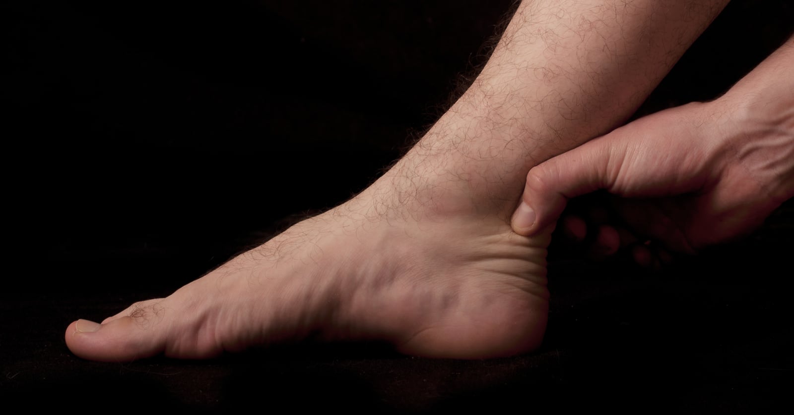 Moody photo of a man’s lower leg and foot on a dark background, with a hand pinching or massaging the achilles tendon.