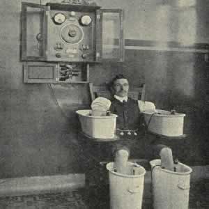 Vintage photo of a man in galvanic bath, seated on a chair between a pair of basins for his legs and a pair for his arms. There’s some old timey electrical hardware on the wall above him.