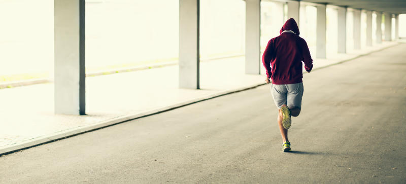 Photo of a male runner with knee pain, jogging away from the camera in a brightly sunlit empty garage.