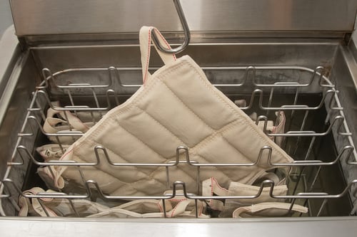Photo of the interior of a hydrocollator tank, showing one steam pack being pulled out the hot water by a metal hook. The sides of the tank are steel or aluminum, and there are several vertical racks in the hot water that resemble oven racks.