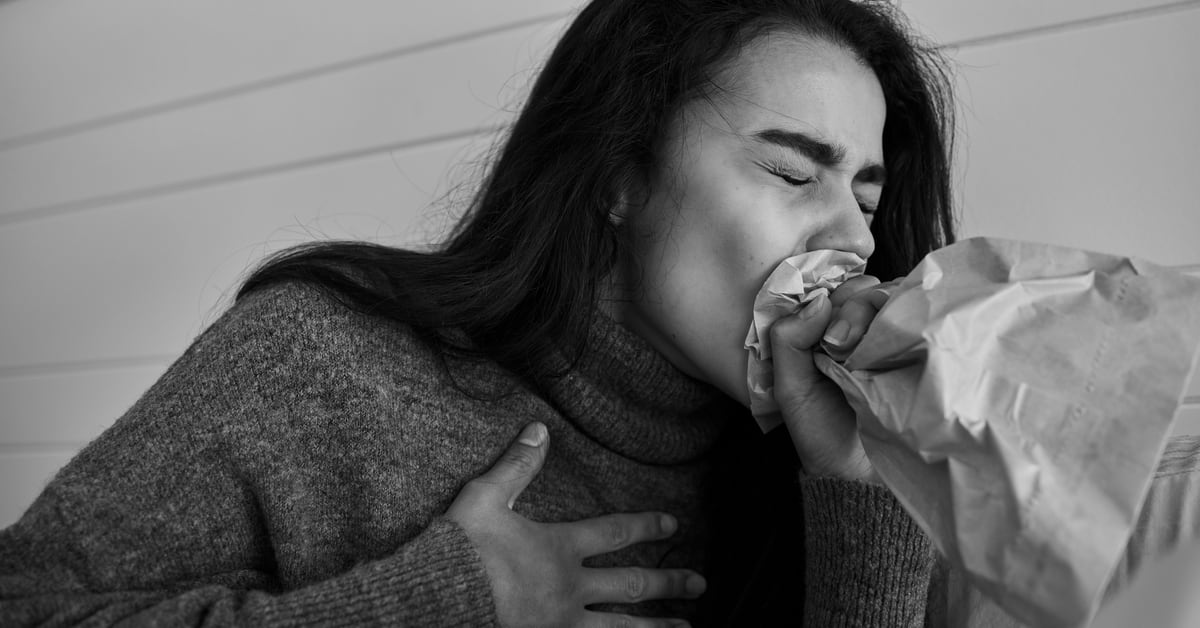 Photo of an Asian woman clutching her chest and breathing into a paper bag, dramatization of a panic attack and hyperventilation. The tone is a bit bleak.