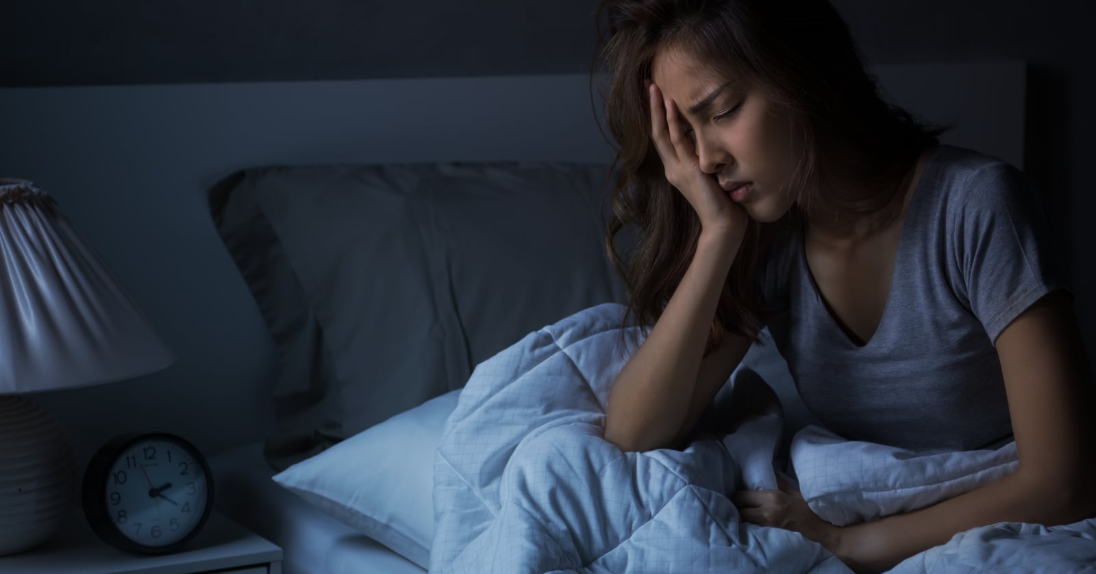 Photo of a young Asian woman sitting in bed in the semi-dark resting her head on one hand, eyes closed.