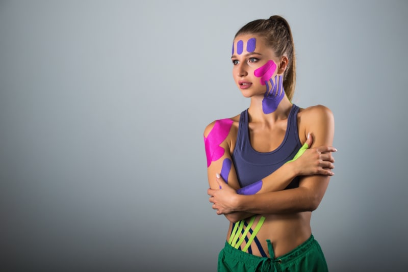 Studio photo of a pretty young female athlete, festooned with a wide variety of colourful therapeutic tapes.