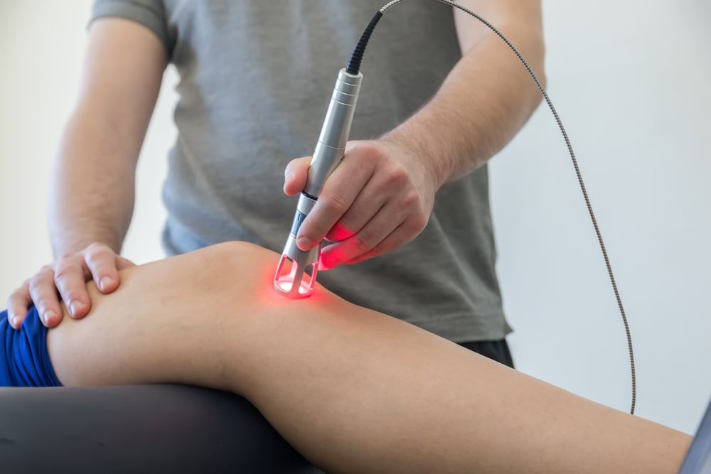 Photo of a therapy session, with a man in a grey shirt applying a technological "wand" with a glowing red tip to the knee of a patient lying on a table in front of him.