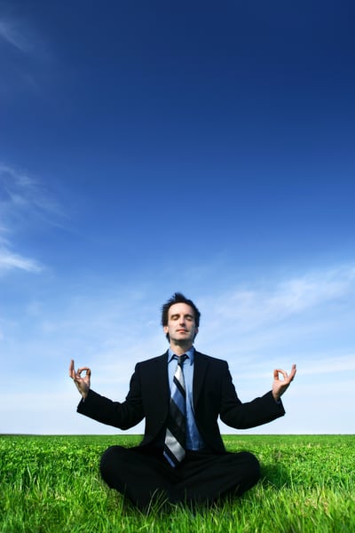 Stereotypical stock photo of a guy in a suit meditating, sitting cross-legged in a field with a blue sky background, representing the common belief back pain might be helped by meditating and/or yoga and stress relief in general.