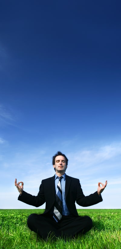 Stereotypical stock photo of a guy in a suit meditating, sitting cross-legged in a field with a blue sky background, representing the common belief back pain might be helped by meditating and/or yoga and stress relief in general.