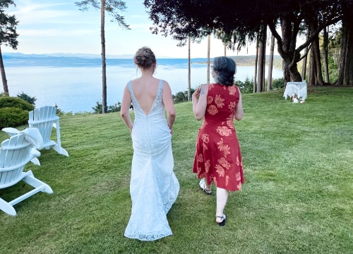 Photo of the bride and my wife facing away, walking on a lush lawn towards a drop-off overlooking trees and a large ocean view.