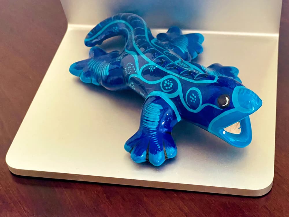The image shows a blue ceramic figurine in the shape of a salamander, decorated with light blue patterns, which include lines and dots that accentuate its body and limbs. The salamander’s open mouth adds character, making it appear as if it’s mid-motion. The glossy, translucent surface suggests it’s crafted from a smooth, polished material. The salamander is placed on an aluminum platform, the stand of an Apple Studio Display on a dark wood desk, contrasting its vibrant blue against a neutral background.