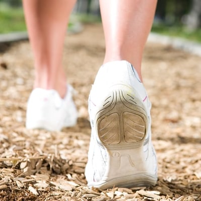 Stock photo of a close-up of a person’s lower legs and feet and white shoes as they walk on a trail covered with wood chips. The background is blurred but appears bright and green, and the setting is probably a park.