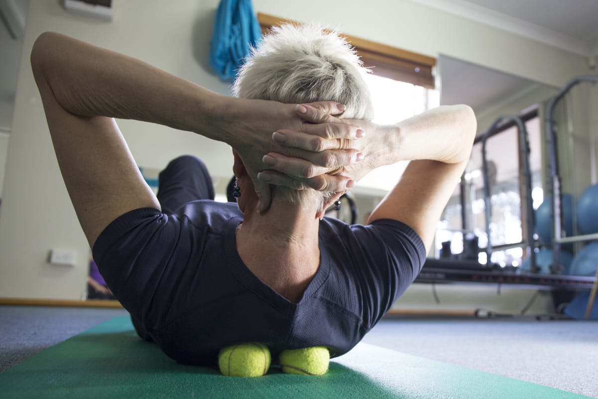 Photo of an older woman in a gym, lying on an exercise matt with 2 tennis balls under her upper back on either side of her spine, with her hands behind her head and slightly flexed upwards like she’s starting a sit-up.