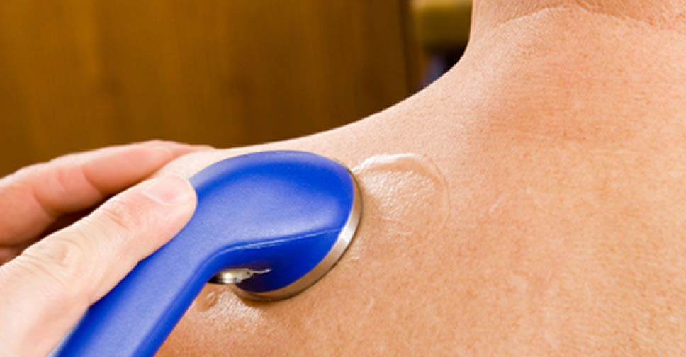 Photo of the left side of a man’s upper back, shoulder, and neck, with a bright blue ultrasound wand being applied to the back of his shoulder.
