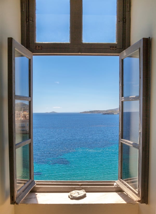 Photo of an open window, looking out onto a view of a sunny, flowery patio with ocean and mountains in the background. It’s bright outside and dark inside.
