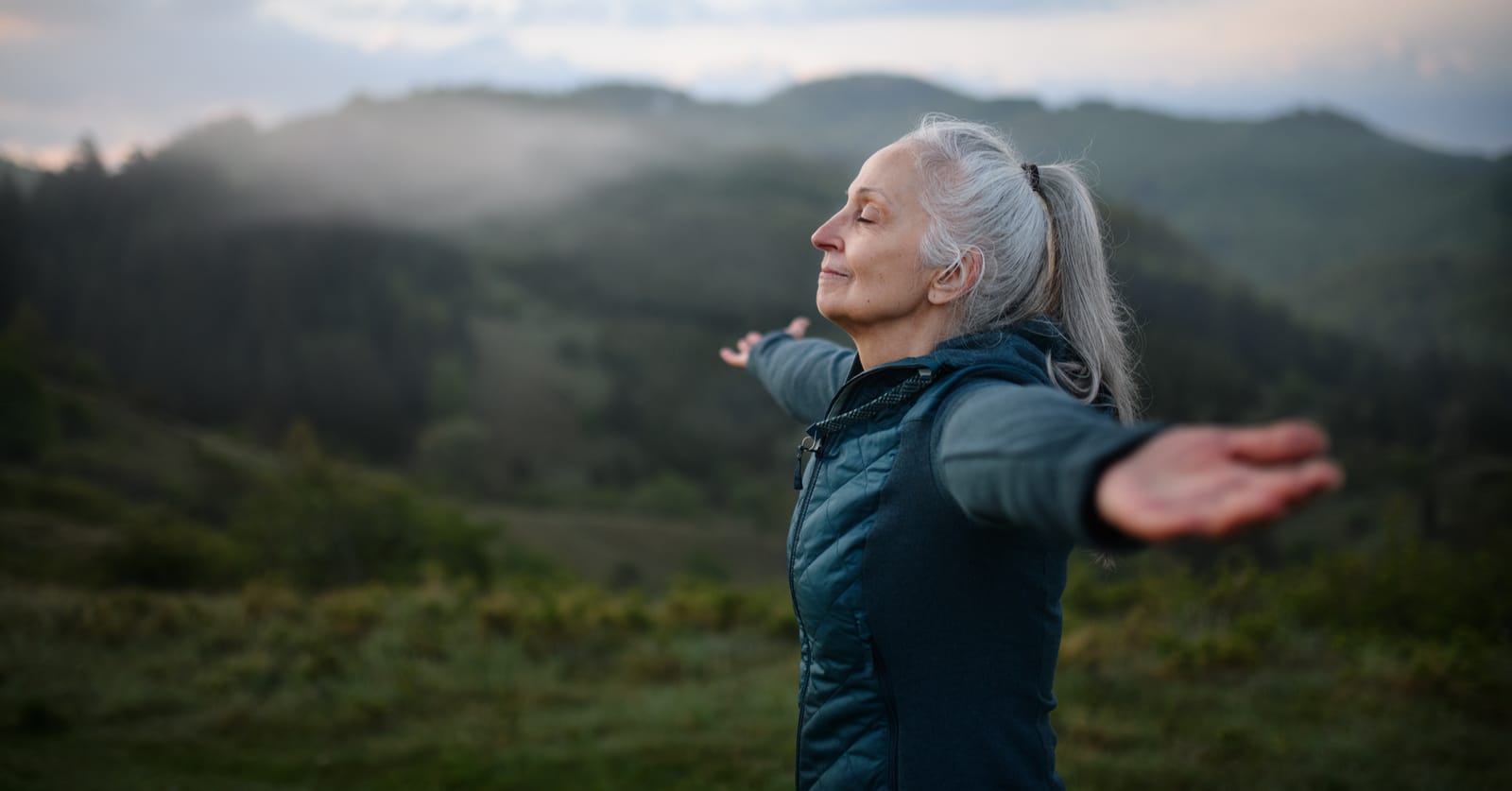Photo of an older woman doing a breathing exercise with a scenic backdrop. Her arms are raised, eyes closed, and head tilted up.