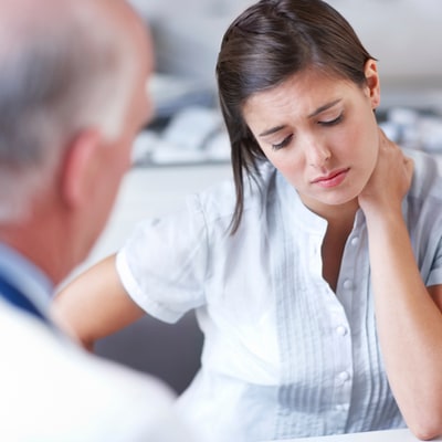 Photo of a female patient holding her neck and talking to a older male doctor in the foreground.