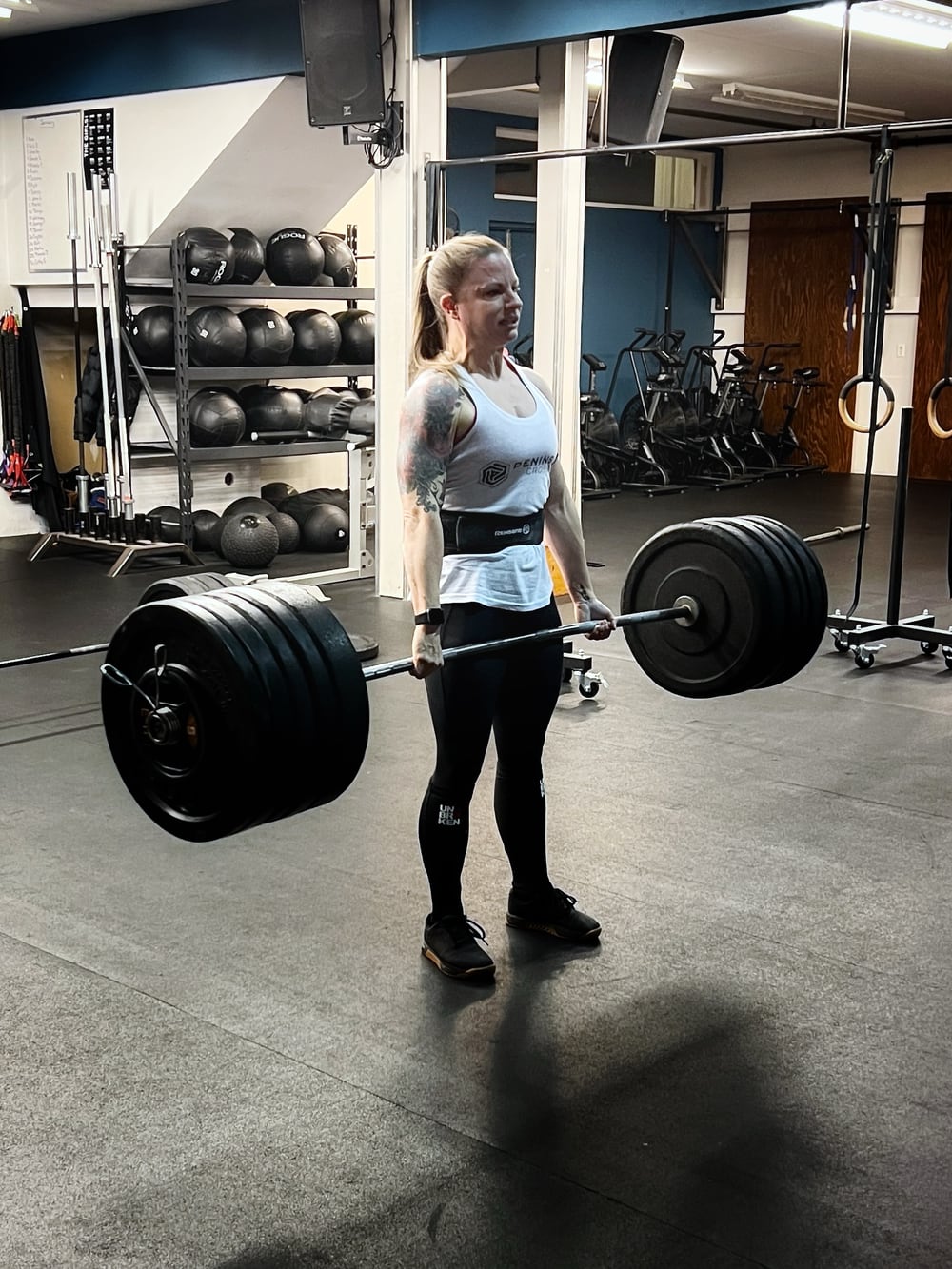 Photo of a woman, Chrystal Ladouceur, in a gym with many spin bikes and medicine balls in the background. She’s lifting an extremely heavy barbell with five huge plates on each end. She’s standing up straight and holding the barbell in front of her at waist level. She’s a blonde with a pony tail, wearing a white tank top, a black weight belt, and black tights, and she has a large, elaborate tattoo of vines and flowers (perhaps) on her right upper arm and shoulder. Her expression is close to a grimace and looks very intense.