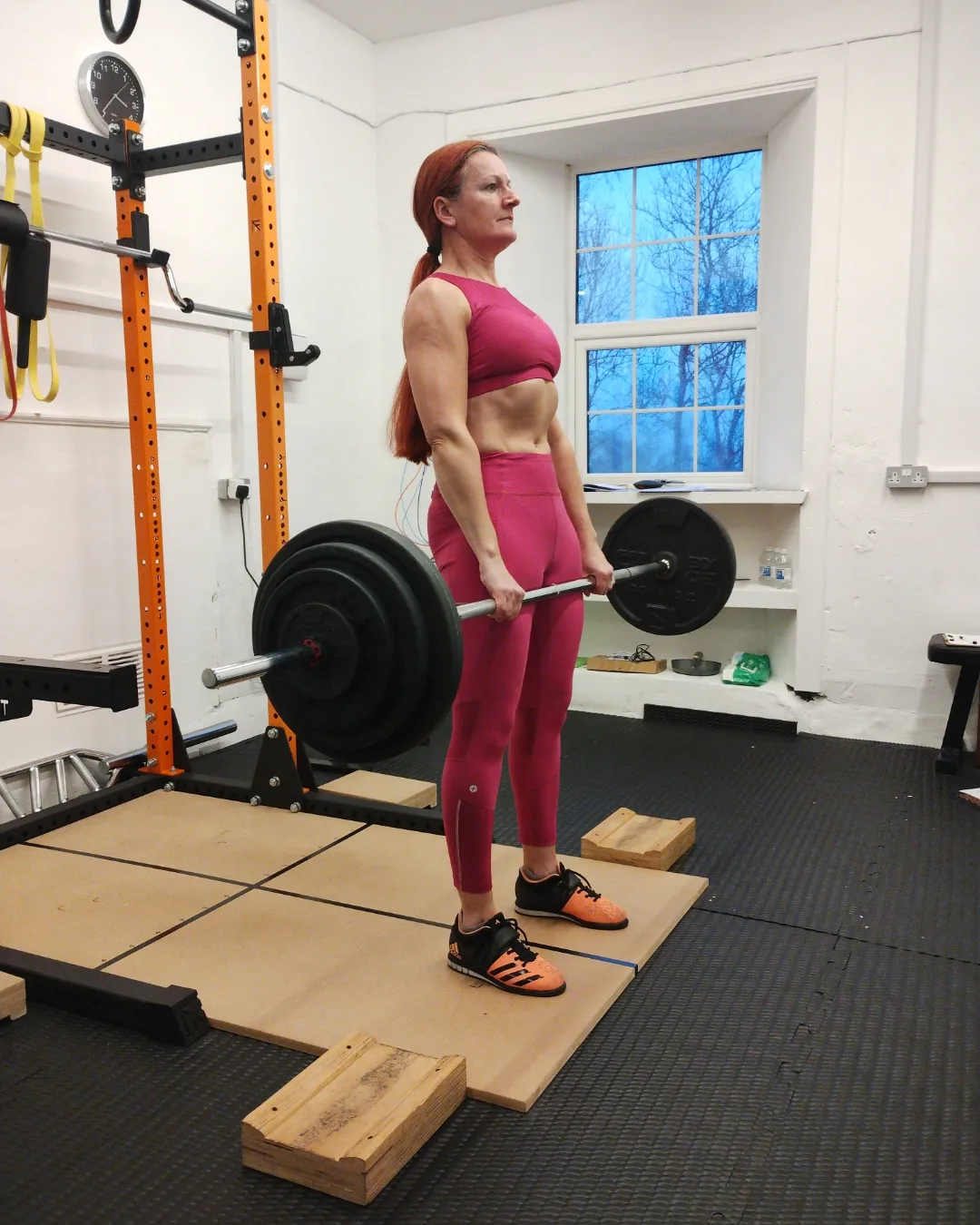 Photo of a woman, Elaine Robertson, lifting a heavy barbell in her home gym, a small white room with a small window in the background looking out onto a cold blue sky and bare trees. She’s standing on a wooden platform, with a bright orange metal lifting rack behind her, holding a barbell that looks like it probably weighs about 60kg. She’s a fit-looking middle-aged women with bright red long hair in a ponytail wearing matching hot pink sports bra and tights.