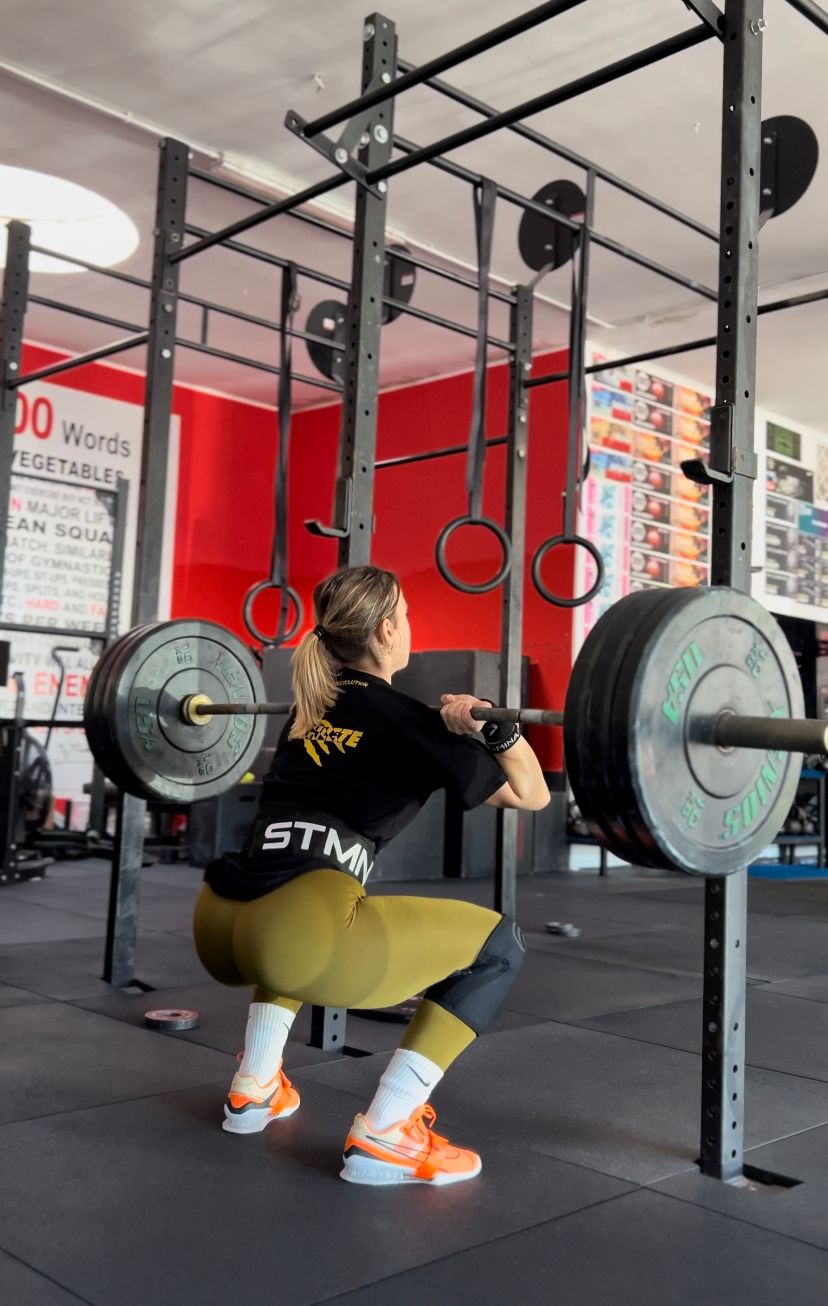 Photo of a woman, Giusi Protopapa, performing a back squat in a gym. She is facing away from the camera, squatting deeply with a loaded barbell with 60kg of plates positioned at her upper chest, gripping the bar with her hands. She is wearing a black shirt, olive-green leggings, and bright orange athletic shoes. The squat rack and gym equipment, including gymnastics rings and pull-up bars, are visible in the background. The lifter appears focused and is wearing a lifting belt for support.