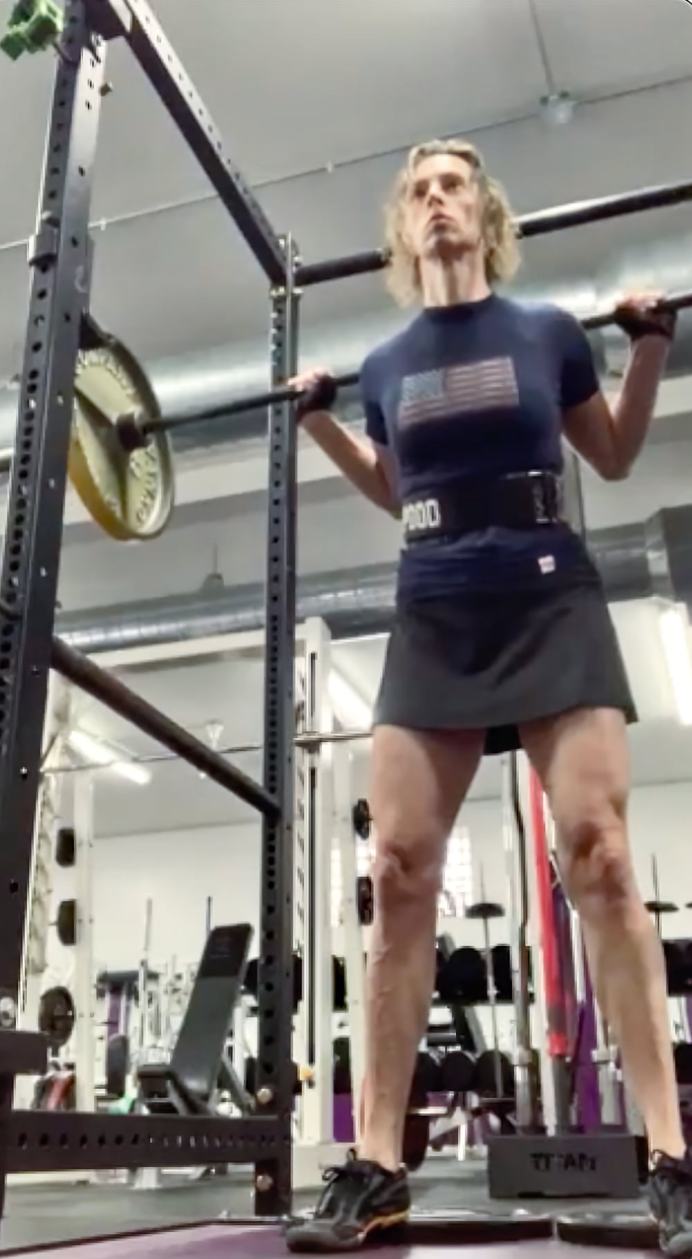 Photo looking up at a woman, Jennifer Adams, in a squat rack in a gym performing a barbell back squat. She appears focused as she braces herself for a squat. She is standing upright with a barbell (loaded with about 20kg of plates) resting across her upper back and holding it securely with her hands. She is wearing a navy blue shirt with an American flag design, a lifting belt, and a black skirt, along with black athletic shoes. The squat rack and other gym equipment are visible in the background.