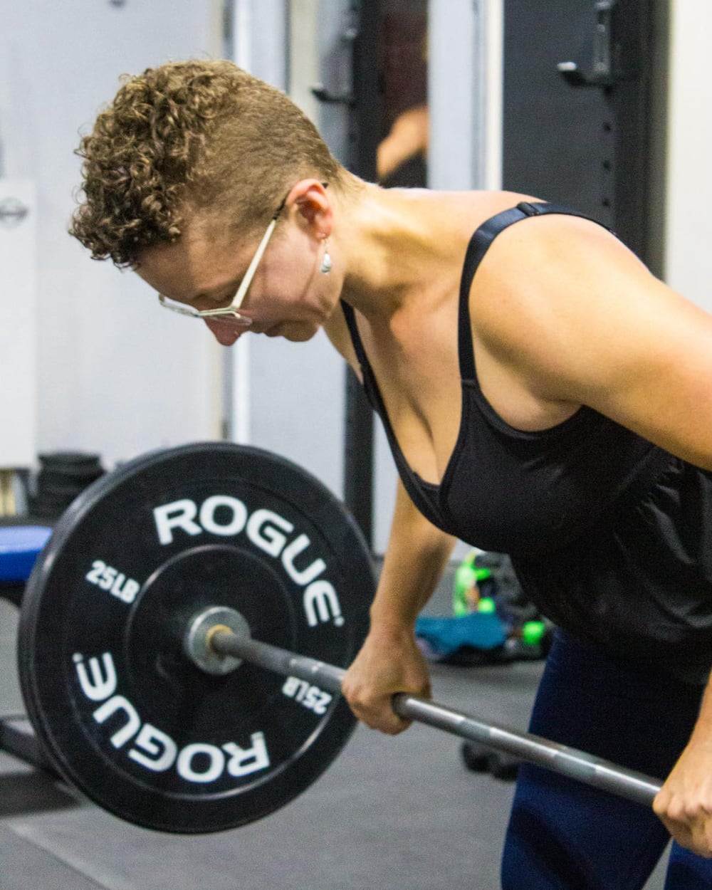 Closeup photo of a woman, Jesse Robson, lifting a barbell with fifty pounds of plates. The photo is cropped mostly to her upper body; she is leaning forward and pulling the barbell up toward her body. She has short curly blonde hair, and she is wearing glasses, a sleeveless black top, and dark athletic pants. Her expression shows focus and determination as she engages her upper back and arms for the exercise. Gym equipment in the background.