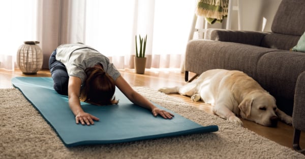 Photograph of a woman in “child’s pose” in her living room. Right beside her, a golden lab is in a nearly identical pose, looking bored and sleepy.